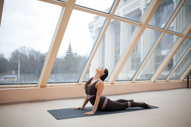 istock brunette woman practicing yoga concept, stretching in Cobra exercise, Bhujangasana pose in front of large windows. working out, full length, wearing sportswear. 931227260