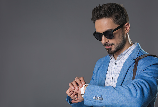 Wait up portrait of handsome calm man in sunglasses checking time at wristwatch. Isolated on gray background. Copy space in left side