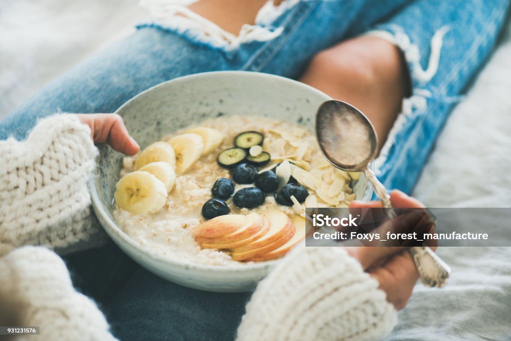 Woman in jeans and sweater eating healthy oatmeal porriage Healthy winter breakfast in bed. Woman in woolen sweater and shabby jeans eating vegan almond milk oatmeal porridge in bowl with berries, fruit and almonds. Clean eating, vegetarian food concept Breakfast Stock Photo