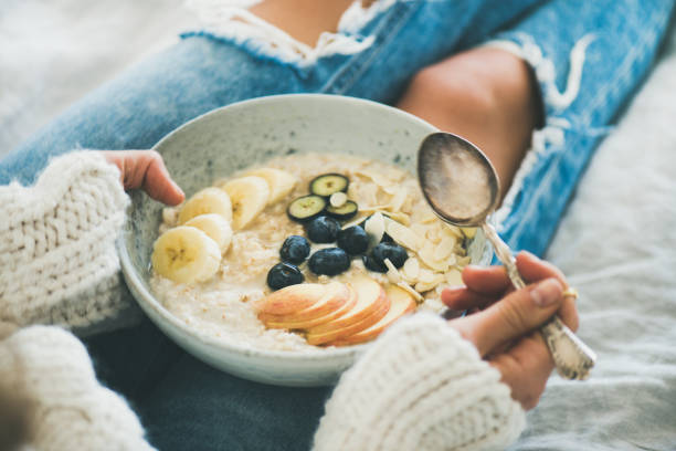 Woman in jeans and sweater eating healthy oatmeal porriage Healthy winter breakfast in bed. Woman in woolen sweater and shabby jeans eating vegan almond milk oatmeal porridge in bowl with berries, fruit and almonds. Clean eating, vegetarian food concept alkaline stock pictures, royalty-free photos & images