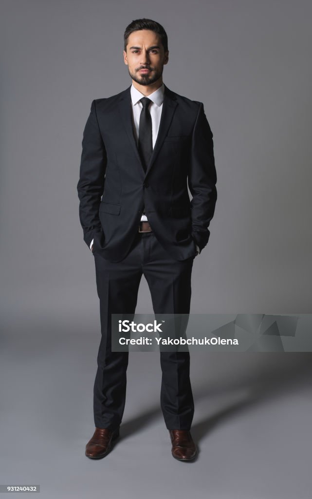Young serious successful businessman standing with hands in pockets Full length portrait of noble concentratad man in suit and posing. Concept of leadership and booming. He is looking at camera with confidence Portrait Stock Photo
