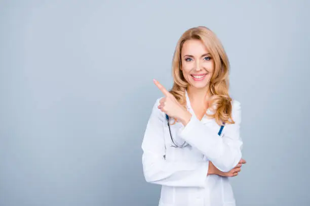 Photo of Look there! Portrait of confident experienced qualified cheerful smiling with blonde hair mature female doc, she is pointing on blank empty space over her shoulder, isolated on grey background