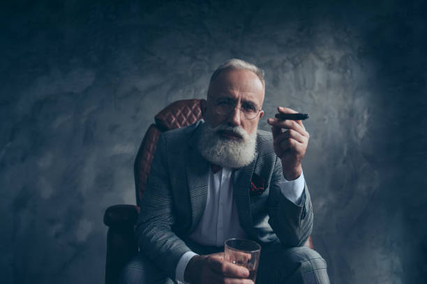 mentalità, meditare uomo ricco in occhiali, tenere sigaretta, bicchiere con brandy, in smoking con papillon rosso, sedersi su sedia di pelle su sfondo grigio, guardando la fotocamera, azioni, azioni, soldi - cigar whisky men smoking foto e immagini stock