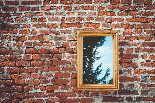 ancient brick wall with golden frame mirror reflecting trees with sky on background