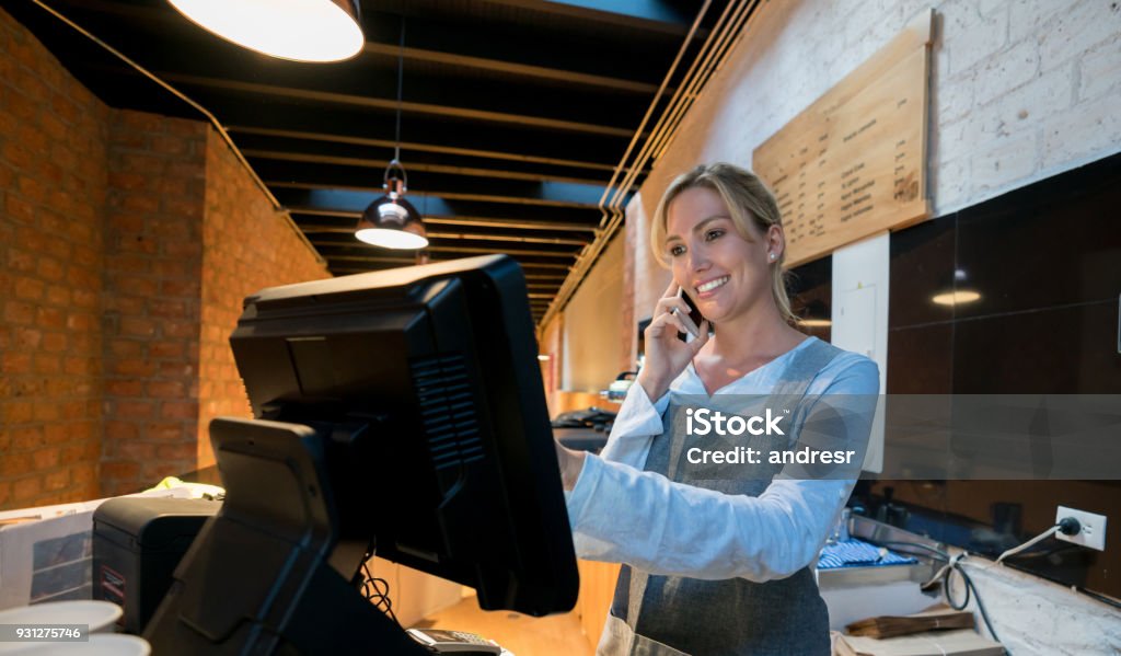 Beautiful saleswoman registering a takeout order while talking with the customer on a smartphone looking happy Beautiful saleswoman registering a takeout order while talking with the customer on a smartphone looking happy and smiling Ordering Stock Photo