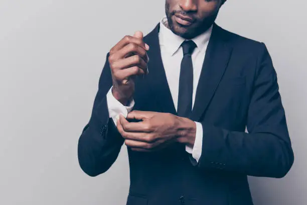 Cropped half face portrait of trendy, attractive, stunning man in black tuxedo with tie fasten button on sleeve cuffs of his white shirt, isolated on grey background