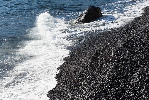 The landscape (or seascape) at a black beach (Greek: Mavra Volia or Maura Volia) consisting of small pebbles of volcanic origin (mostly of black color, but also some white, yellow, red, orange and green) close to the Emporio (or Emporios) village on the Greek Island of Chios. In the background is the Aegean Sea (with waves and surf). The pebbles were created from the lava of Psaronas volcano (now inactive).