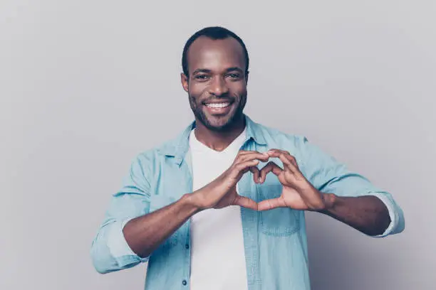 Photo of Portrait of creative, romantic, positive, cheerful, confident guy making love symbol, heart figure with fingers, looking at camera, isolated on grey background