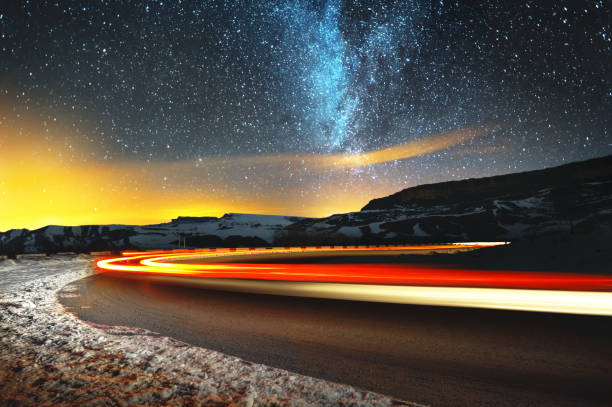 paysage de nuit. ciel de nuit avec un hémisphère nord voie lactée et des étoiles. la route de nuit éclairée par la voiture enroule avec une serpentine et laisse à une distance d’un pied d’un rocher aigu. sentiers de lumière - long exposure rock cloud sky photos et images de collection