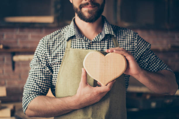 adoro il mio lavoro! ritagliato foto ravvicinata di felice allegro falegname gioioso innamorato, sta mostrando un cuore fatto di legno - carpentry workshop work tool craft foto e immagini stock