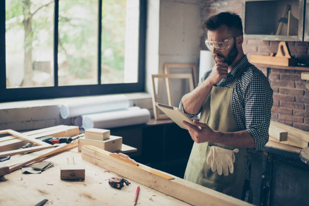 Pensive minded concentrated confident handsome bearded wearing checkered shirt cabinet-maker is watching online videos on the internet how to process wood, he is holding digital tablet