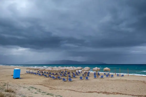 Beach umbrellas and sunbeds on a deserted beach on a rainy day, nearby Rethimnon, Crete, Greece. Akrotiri peninsula visible in the background.