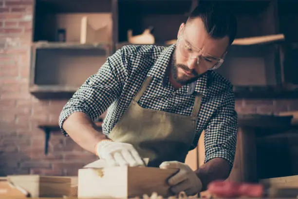 Handsome gifted hardworking confident concentrated bearded handyman clothed in checkered shirt apron gloves and safety glasses is sanding a little wooden casket with emery paper, working in a garage
