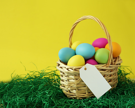 Colorful Easter eggs basket on green grass greeting card with white blank paper label tag for message and copy space on yellow background.