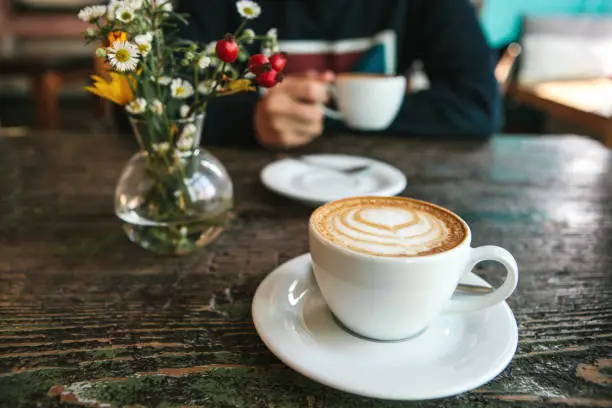Photo of Two cups of coffee and a vase of flowers on a wooden table, the man holds in her hand one cup of coffee in the background. A photo indicates a meeting of people and a joint pastime.