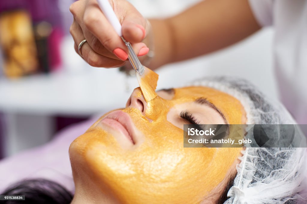 Macrophotographie de la main de l’esthéticienne à l’aide d’une brosse spéciale met sur le visage de la jeune fille un masque d’or dans le spa - Photo de Or - Métal libre de droits