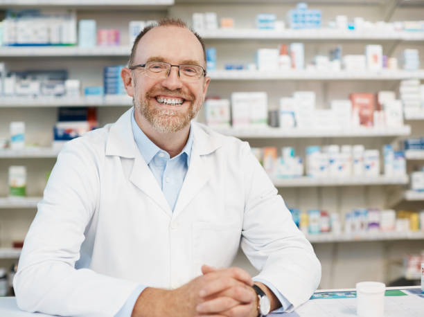 souriant joyeusement pharmacien mâle debout derrière le comptoir de l’officine - pill bottle pharmacy medicine shelf photos et images de collection