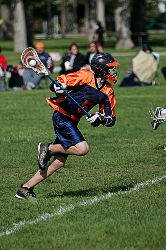 A royalty free DSLR action photo of a lacrosse player in full sprint attack with the ball clearly visible in the pocket of his lacrosse stick. The player is dressed in orange and dark blue surrounded by a field of green turf with spectators looking on from the sidelines. This action image was captured in actual game conditions reflecting competitive intensity.