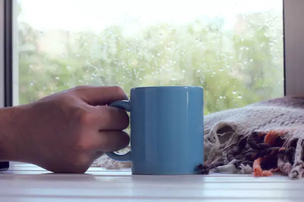 Big blue mug in hand against the window with rain drops