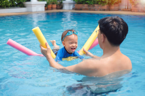 cute little asian 18 months / 1 year old toddler boy child wear swimming goggles learning to swim with pool noodle at outdoor pool; dad and son relaxing in swimming pool of clubhouse in summer day - swimming child swimwear little boys imagens e fotografias de stock