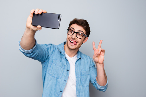 Portrait of positive, comic, trendy guy with stubble shooting selfie on smart phone, using gadget device, gesture v-sign, having video-call, isolated on grey background