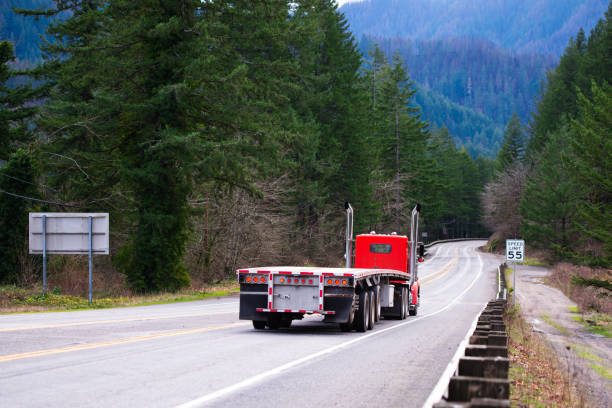 Red big rig semi truck tractor going with empty flat bed semi trailer on the awesome winding road in green forest in Columbia Gorge area