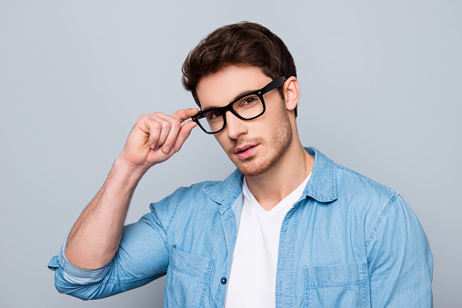 Portrait of stunning, brutal, sexy, concentrated, cool man in jeans shirt holding eyelet of glasses on his face with fingers, looking at camera, isolated on grey background