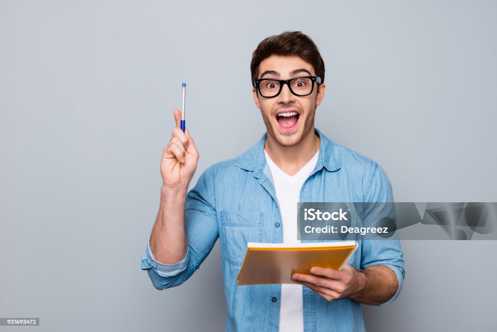 Un mec beau, attrayant, heureux, positif, drôle dans des verres avec la bouche grande ouverte a finalement trouvé une solution comment faire l’exercice, avoir soulevé stylo et carnet en mains, isolé sur fond gris - Photo de Hommes libre de droits