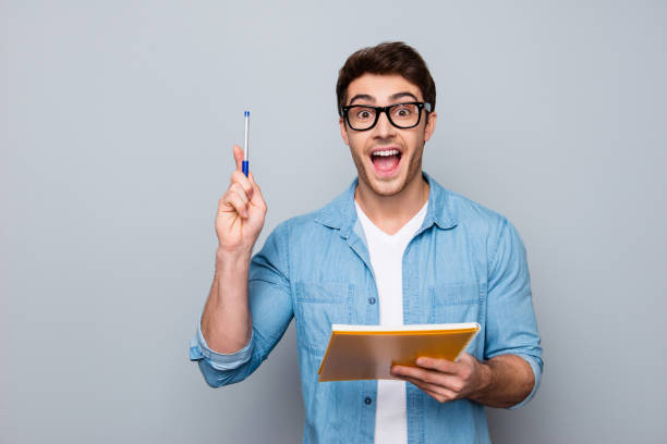 handsome, attractive, glad, positive, funny guy in glasses with wide open mouth finally found a solution how to make exercise, having raised pen and notebook in hands, isolated on grey background - look for trains” imagens e fotografias de stock