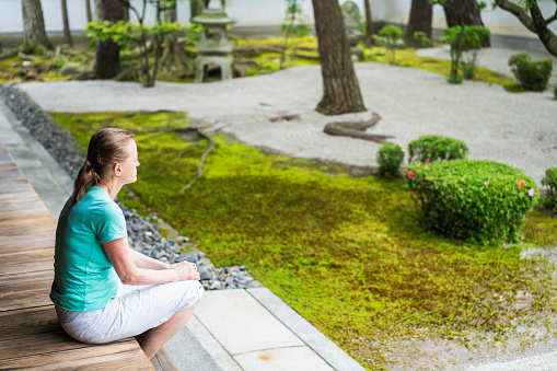 Side view ofseniior woman sitting on temple podium, while enjoying Zen garden in between Chion-ji Temple buildings walls. It is Japanese type rock garden presenting miniature landscape. It is carefully arranged meditation place with rocks, water, or water features, sand, moss, trees and bushes.