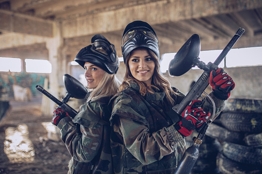 Two women, playing paintball on a rural terrain in abandoned building, beautiful girls holding guns