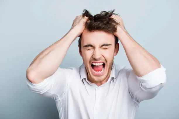Photo of Emotions, stress, madness and people concept - crazy shouting man rending his hair in white shirt, screaming with close eyes and wide open mouth, holding hands on head over gray background