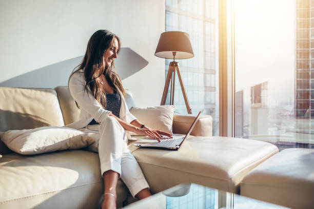 mulher de negócios de fêmea jovem elegante usando um laptop sentado num sofá em casa - luxury apartment - fotografias e filmes do acervo