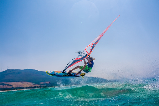 Young man jumping on waves with colourful windsurfing board and sail.