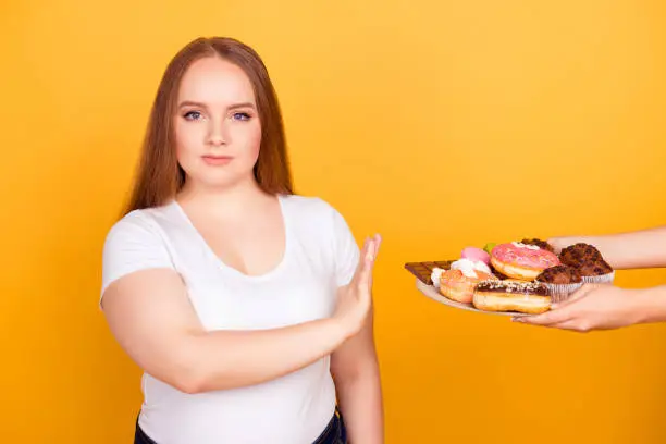 Photo of I'm against eating products containing fat! Will-powered woman wearing white tshirt is refusing to consume tasty delicious sweets on a plate, isolated on bright yellow background