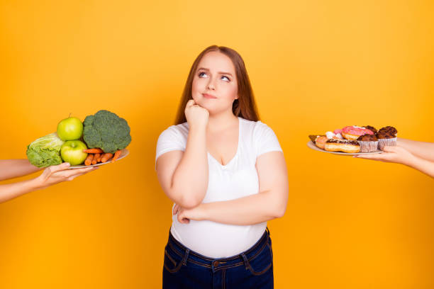 body care bodycare concept. confused thoughtful hesitating fatty woman can't make a choice between healthy natural products and sugary tasty biscuits, isolated on bright yellow background - dieting overweight weight scale healthcare and medicine imagens e fotografias de stock