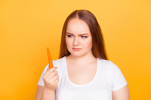 I want to be slim I need to eat fresh food! Close up portrait of unhappy sad and unsatisfied fatty woman wearing white tshirt, she is looking at carrot in her hand isolated on bright yellow background