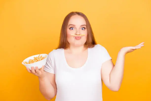 Bodycare body care concept. Where is the french fries? Happy wondered cheerful fatty lady is fooling around, she is making fake mustache using fried potatoes, isolated on bright yellow background