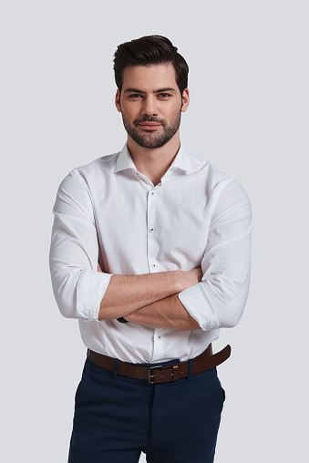 Good looking young man looking at camera with smile and keeping arms crossed while standing against grey background