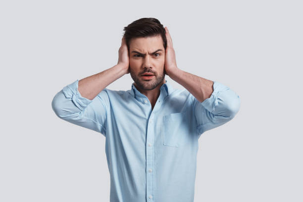 Too loud. Frustrated young man covering ears with hands and looking at camera while standing grey background hands covering ears stock pictures, royalty-free photos & images