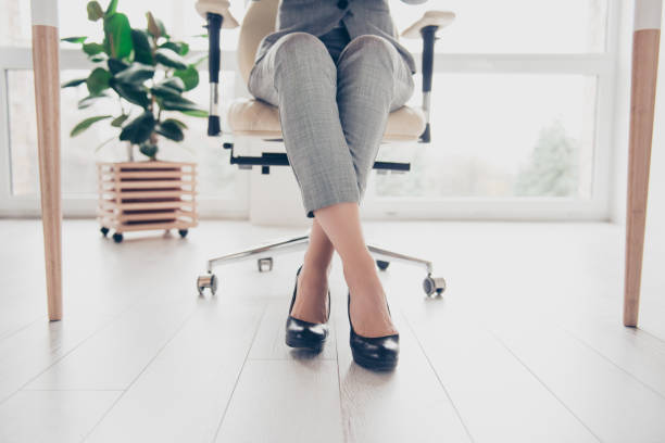 cropped close up photo of healthy beautiful elegant woman's legs wearing high-heeled shiny black shoes, the woman is sitting in office at the table on modern luxurious armchair - fato de senhora imagens e fotografias de stock