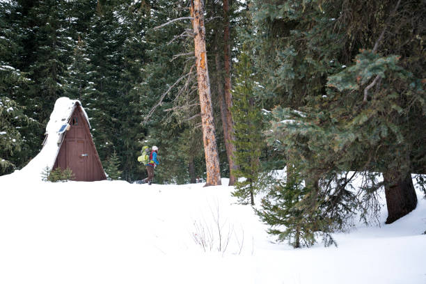 Young man in 20's backpacking in a snowy forest walks past a triangular wooden hut, Hyalite Canyon Recreational Area, Bozeman, Montana, USA