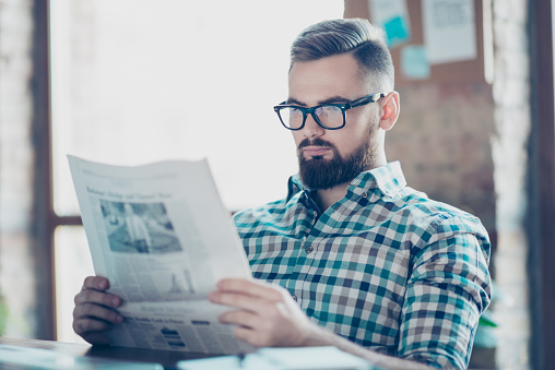 Portrait of serious confident concentrated young stylish guy wearing glasses and checkered shirt, he is reading a newspaper, getting knowledge about current events