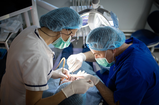 Two dental doctors, man and woman, examining patient on dentist chair and doing surgery. They wear protective mask, gloves and caps. Real people, real surgery in this series