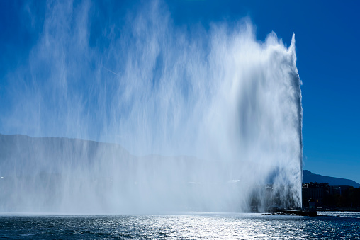 In the morning, the Jet d'Eau in the harbor of Geneva with the lake, the city and its old town. The west wind causes a large plume that is highlighted by the sun.