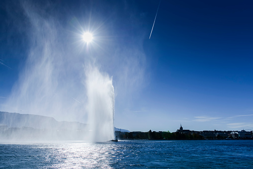 In the morning, the Jet d'Eau in the harbor of Geneva with the lake, the city and its old town. The west wind causes a large plume that is highlighted by the sun.