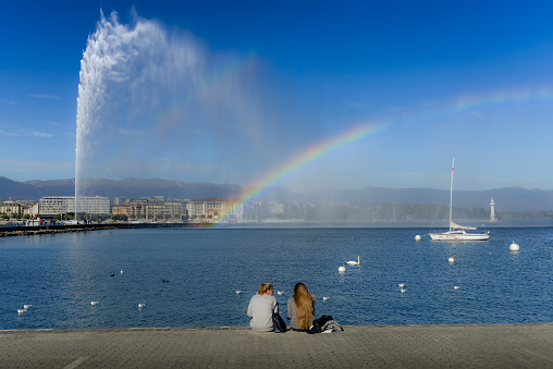 In the early morning, the Jet d'Eau in the harbor of Geneva with the lake, the city and its palaces. The west wind causes a large plume and a rainbow.