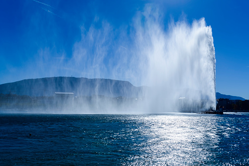 In the morning, the Jet d'Eau in the harbor of Geneva with the lake, the city and its old town. The west wind causes a large plume that is highlighted by the sun.