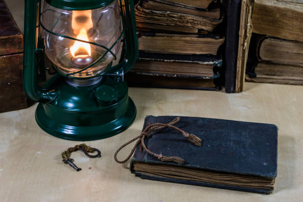 istock Old oil lamp and books lying on the table. A flame from an old source of light illuminating old prints. Black background. 931823614