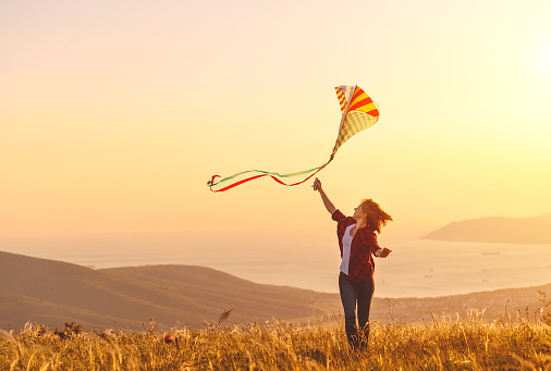 Happy young woman running with a kite on a glade at sunset in summer